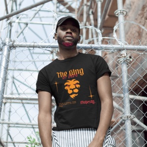 Un bărbat poartă un tricou negru cu textul 'The King is here' și o grafică a unui leu stilizat în portocaliu. El are o barbă colorată parțial în roșu, o șapcă neagră și stă în fața unui gard metalic cu sârmă ghimpată, cu o structură industrială pe fundal.