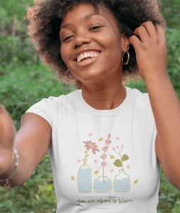 Tânără zâmbitoare purtând un tricou alb cu design floral care ilustrează trei flori delicate în vaze și mesajul 'you are about to bloom'. Fata are părul creț și poartă cercei rotunzi, iar fundalul verde sugerează un mediu natural și relaxat.