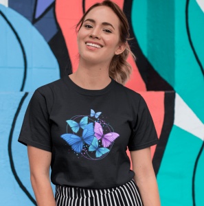 Fată zâmbitoare purtând un tricou negru cu un design grafic de fluturi albastri și mov, pozând în fața unui mural colorat.