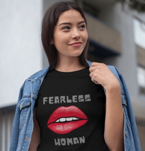 Fată purtând un tricou negru cu designul 'Fearless Woman' și buze roșii grafice, având o jachetă de blugi pe umeri și zâmbind într-un cadru urban.