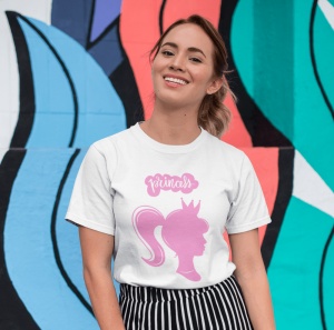 Tânără purtând un tricou alb cu un design roz care înfățișează silueta unei prințese cu o coroană și cuvântul 'princess' deasupra. Ea zâmbește în fața unui perete viu colorat, decorat cu forme abstracte în nuanțe de albastru, verde și portocaliu.