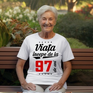 O femeie purtând un tricou alb cu textul  'Viața începe de la 97 de ani'. Fundal din lemn cu diferite nuanțe de maro și verde.