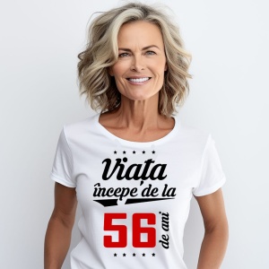 O femeie purtând un tricou alb cu textul  'Viața începe de la 56 de ani'. Fundal din lemn cu diferite nuanțe de maro și verde.