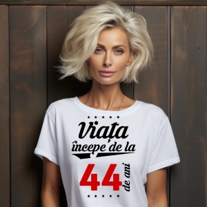 O femeie purtând un tricou alb cu textul  'Viața începe de la 44 de ani'. Fundal din lemn cu diferite nuanțe de maro și verde.
