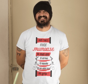 Bărbat zâmbitor, cu barbă și pălărie neagră, purtând un tricou alb cu mesajul 'Am trei fiice frumoase și mai am o armă, o lopată și un alibi bun. Ai grijă!', decorat cu ilustrații de arme, lopată și cătușe. Bărbatul are un tatuaj pe brațul drept și stă pe