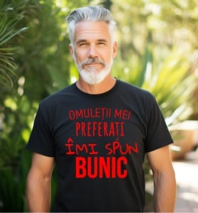 Un bărbat matur cu barbă albă poartă un tricou negru inscripționat cu text roșu: "Omuleții mei preferați îmi spun BUNIC". În fundal, se observă un peisaj verde, sugerând o zi însorită în natură.