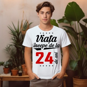 Un tânăr stând într-o cameră cu plante, purtând un tricou alb cu mesajul: „Viața începe de la 24 de ani”. Tânărul este în fața unui fundal cu plante și obiecte de decor.