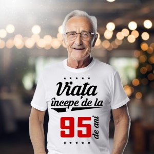 Bărbat în vârstă purtând ochelari și un tricou alb cu mesajul 'Viața începe de la 95 de ani', într-un cadru iluminat cald.