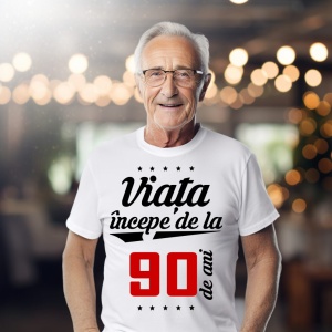 Bărbat în vârstă purtând ochelari și un tricou alb cu mesajul 'Viața începe de la 90 de ani', într-un cadru iluminat cald.