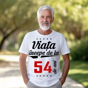 Bărbat în vârstă cu barbă albă, zâmbitor, purtând un tricou alb cu mesajul 'Viața începe de la 54 de ani', stând într-un parc luminat de soare.