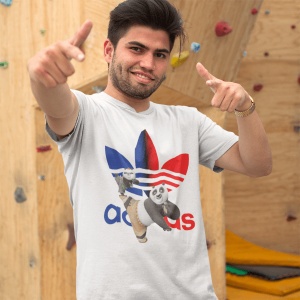 Bărbat zâmbitor purtând un tricou alb cu un design colorat, ce include personaje animate(KungFu Panda Po) și un logo reinterpretat, gesticulând energic într-un spațiu interior decorat cu panouri de escaladă.