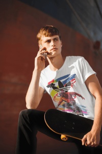 Tânăr purtând un tricou alb cu un design vibrant, reprezentând un skater și un fundal abstract colorat, vorbind la telefon și ținând un skateboard, în fața unui perete portocaliu.