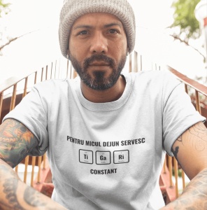 Bărbat purtând o căciulă gri și un tricou alb cu textul 'PENTRU MICUL DEJUN SERVESC Ti Ga Ri CONSTANT,' stilizat ca elemente chimice din tabelul periodic. Bărbatul are tatuaje pe brațe și este fotografiat în aer liber, sprijinit de o scară sau balustradă 