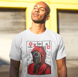 Un bărbat purtând un tricou alb cu un design inspirat din Squid Game, înfățișând un personaj cu mască roșie și simboluri geometrice, pozând în lumina soarelui în fața unui fundal galben.