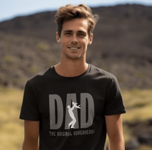 Bărbat zâmbitor purtând un tricou negru cu mesajul 'DAD - The Original Superhero!' și o siluetă de tată care ridică un copil, încorporată în literele mari ale cuvântului 'DAD.' Fundalul arată un peisaj natural cu dealuri stâncoase.