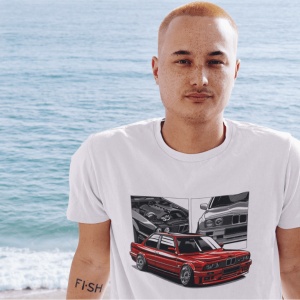 Bărbat purtând un tricou alb cu o ilustrație grafică detaliată a unui BMW roșu clasic, pozat pe fundalul unei plaje cu oceanul în fundal.