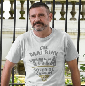Un bărbat poartă un tricou alb cu mesajul 'Cel mai bun tată șofer de camion'. Fundalul include un gard de fier forjat și elemente arhitecturale clasice.