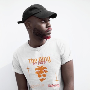 Un bărbat poartă un tricou alb cu inscripția 'The King is here' și o grafică a unui leu stilizat în portocaliu. El are o șapcă neagră și barba parțial colorată în roșu, privind lateral. Fundalul este simplu, alb.
