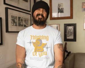 Un bărbat cu barbă și căciulă neagră zâmbește în timp ce poartă un tricou alb cu un design vintage. Designul conține un înger stilizat și textul 'Thinking of You' scris într-un font gotic galben. Sub imaginea îngerului apare un mesaj mic de afecțiune. Băr