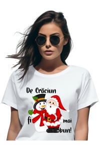 Femeie purtând ochelari de soare și un tricou alb cu un design ce îl ilustrează pe Moș Crăciun și un om de zăpadă ținându-se de mână, alături de textul 'De Crăciun fii mai bun!'