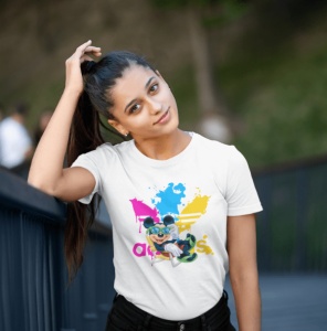 Femeie purtând un tricou alb cu un design colorat ce include logo-ul 'Adidas' și un personaj stilizat(Mickey Mouse), stând afară și ținându-se cu o mână de părul prins în coadă de cal, cu o expresie relaxată.