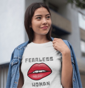 Fată purtând un tricou alb cu designul 'Fearless Woman' și buze roșii grafice, având o jachetă de blugi pe umeri și zâmbind într-un cadru urban.