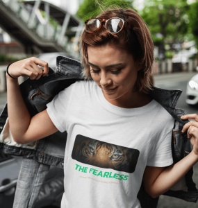 Fată purtând un tricou alb cu un design grafic ce prezintă ochii unui tigru și textul 'THE FEARLESS', scoțându-și jacheta de blugi, într-un mediu urban.