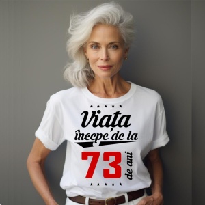 O femeie purtând un tricou alb cu textul  'Viața începe de la 73 de ani'. Fundal din lemn cu diferite nuanțe de maro și verde.