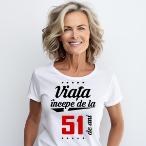 O femeie purtând un tricou alb cu textul  'Viața începe de la 51 de ani'. Fundal din lemn cu diferite nuanțe de maro și verde.