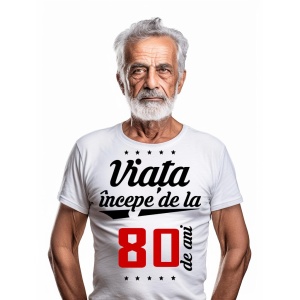 Bărbat în vârstă cu barbă albă, purtând un tricou alb cu mesajul 'Viața începe de la 80 de ani', pe un fundal alb simplu.