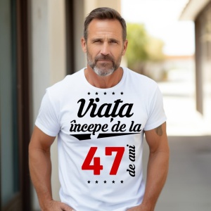 Bărbat matur cu barbă scurtă, purtând un tricou alb cu mesajul 'Viața începe de la 47 de ani', stând în exterior pe o alee însorită.