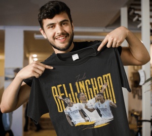 Un bărbat zâmbitor ține în mână un tricou negru cu un design grafic dedicat jucătorului de fotbal Jude Bellingham, incluzând imagini ale acestuia în acțiune și numele său scris cu litere galbene mari.