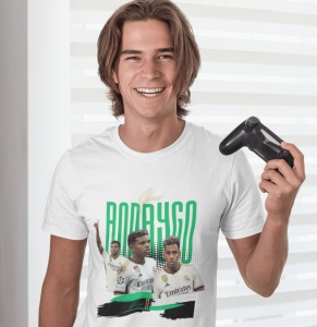 Un tânăr zâmbitor, ținând un controller de joc video, poartă un tricou alb cu design grafic dedicat jucătorului de fotbal Rodrygo, cu imagini ale acestuia și numele său scris cu litere verzi.
