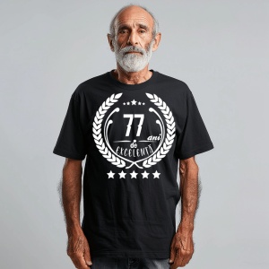 Un bărbat zâmbitor purtând un tricou negru cu textul ''77 de ani de excelență'', înconjurat de o coroană de lauri.