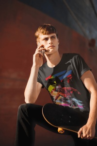 Tânăr purtând un tricou negru cu un design vibrant, reprezentând un skater și un fundal abstract colorat, vorbind la telefon și ținând un skateboard, în fața unui perete portocaliu.