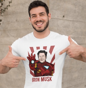 Bărbat zâmbitor și cu barbă, purtând un tricou alb cu un design amuzant care îl arată pe Elon Musk în costumul lui Iron Man, cu textul 'IRON MUSK' dedesubt. Bărbatul indică cu degetele spre designul tricoului, pe un fundal de perete texturat.