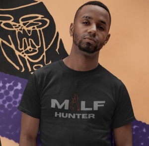 Un bărbat poartă un tricou negru cu textul 'MILF HUNTER' în font gri și o ilustrație stilizată în roșu. Fundalul este un perete pictat cu un design artistic abstract.