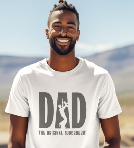 Bărbat zâmbitor purtând un tricou alb cu mesajul 'DAD - The Original Superhero!' și o siluetă de tată care ridică un copil, încorporată în literele mari ale cuvântului 'DAD.' Fundalul arată un peisaj natural cu dealuri și cer senin.