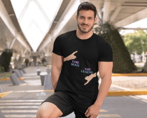 Un bărbat zâmbitor poartă un tricou negru cu un design amuzant ce include două mâini care arată cu degetul, una în sus cu textul 'The Man' și una în jos cu textul 'The Legend'. Bărbatul pozează într-un spațiu urban cu o structură arhitecturală modernă dea