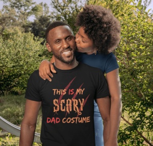 Un bărbat care zâmbește, purtând un tricou negru cu textul 'This is my scary dad costume' cu un design cu zgârieturi de gheare, este îmbrățișat de o femeie care îi dă un sărut pe obraz. Ambii sunt în aer liber, în fața unei scene verzi de parc sau grădină