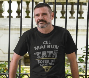 Un bărbat poartă un tricou negru cu mesajul 'Cel mai bun tată șofer de camion'. Fundalul include un gard de fier forjat și elemente arhitecturale clasice.