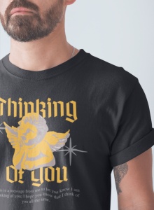 Un bărbat cu barbă poartă un tricou negru cu design retro care afișează un înger stilizat, cu textul 'Thinking of You' în font gotic de culoare galbenă. În partea de jos a designului apare un mesaj mic, care exprimă gânduri de afecțiune. Fundalul este sim