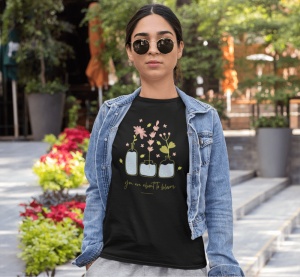 Tânără purtând un tricou negru cu un design floral care ilustrează trei vaze cu flori delicate și mesajul 'you are about to bloom'. Ea poartă o jachetă din denim deschisă și ochelari de soare, stând într-un cadru urban cu vegetație și flori colorate în fu