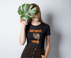 Tânără purtând un tricou negru cu un design care înfățișează un leneș atârnând de o ramură și textul 'JUST DO IT. LATER' scris în portocaliu. Ea ține o frunză mare de monstera în fața feței, lăsând să se vadă doar o parte din aceasta. Fundalul este simplu