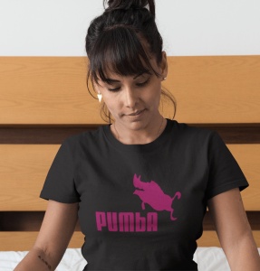 Femeie cu părul prins și breton, purtând cercei în formă de inimă, îmbrăcată într-un tricou negru cu un design roz ce prezintă silueta unui facocer și textul 'PUMBA'. Femeia se uită în jos cu o expresie calmă și relaxată. Fundalul este parțial din lemn, o