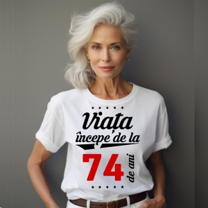 O femeie purtând un tricou alb cu textul  'Viața începe de la 74 de ani'. Fundal din lemn cu diferite nuanțe de maro și verde.