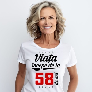 O femeie purtând un tricou alb cu textul  'Viața începe de la 58 de ani'. Fundal din lemn cu diferite nuanțe de maro și verde.