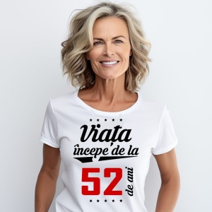 O femeie purtând un tricou alb cu textul  'Viața începe de la 52 de ani'. Fundal din lemn cu diferite nuanțe de maro și verde.