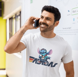 Bărbat zâmbitor vorbind la telefon, purtând un tricou alb cu un design colorat care include un personaj animat albastru(Stitch) și logo-ul Adidas stilizat. Fundalul arată un perete decorativ cu text și grafică.