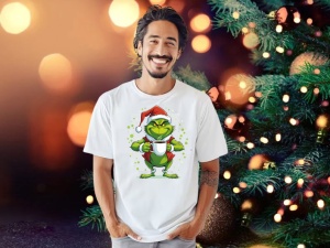 Bărbat zâmbitor purtând un tricou alb cu un desen de Crăciun reprezentând un personaj verde în costumul lui Moș Crăciun, ținând o cană, în fața unui fundal festiv cu lumini strălucitoare și un brad de Crăciun decorat.
